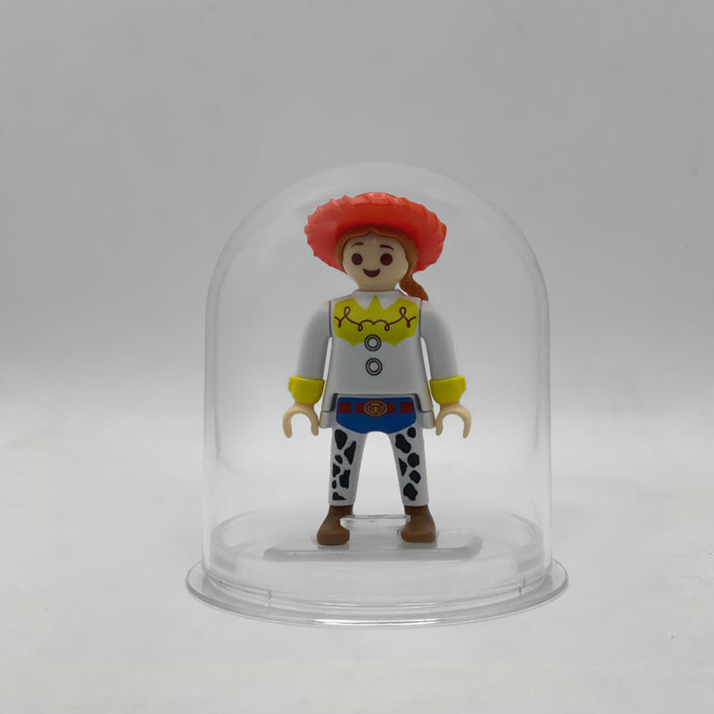 Minifigura Playmobil de Jessie la vaquera con sombrero rojo, trenza pelirroja y zahones de vaca, sobre su peana original Playmobil dentro de la urna expositora transparente, colección Disney Kinder FCO1008