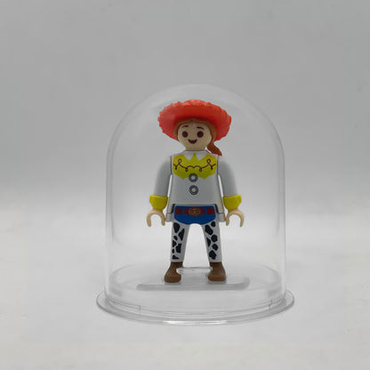Minifigura Playmobil de Jessie la vaquera con sombrero rojo, trenza pelirroja y zahones de vaca, sobre su peana original Playmobil dentro de la urna expositora transparente, colección Disney Kinder FCO1008