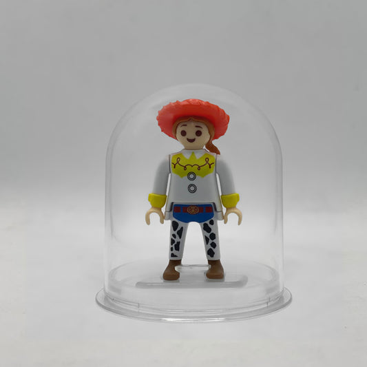 Minifigura Playmobil de Jessie la vaquera con sombrero rojo, trenza pelirroja y zahones de vaca, sobre su peana original Playmobil dentro de la urna expositora transparente, colección Disney Kinder FCO1008