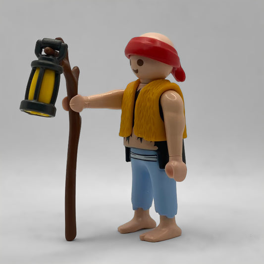 Figura Playmobil FCO5031 de perfil: pañuelo rojo, chaleco ocre de lana, faja de rayas, pantalón azul deshilachado y pies descalzos, con bastón y farol