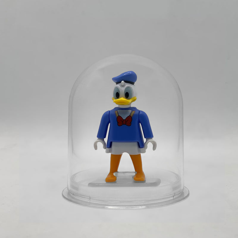 Minifigura Playmobil del Pato Donald con gorra de marinero azul, casaca con pajarita roja y pies naranjas, sobre su peana original Playmobil dentro de la urna expositora transparente, colección Disney Kinder FCO1010