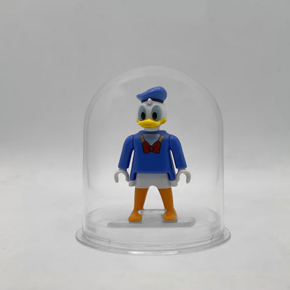 Minifigura Playmobil del Pato Donald con gorra de marinero azul, casaca con pajarita roja y pies naranjas, sobre su peana original Playmobil dentro de la urna expositora transparente, colección Disney Kinder FCO1010