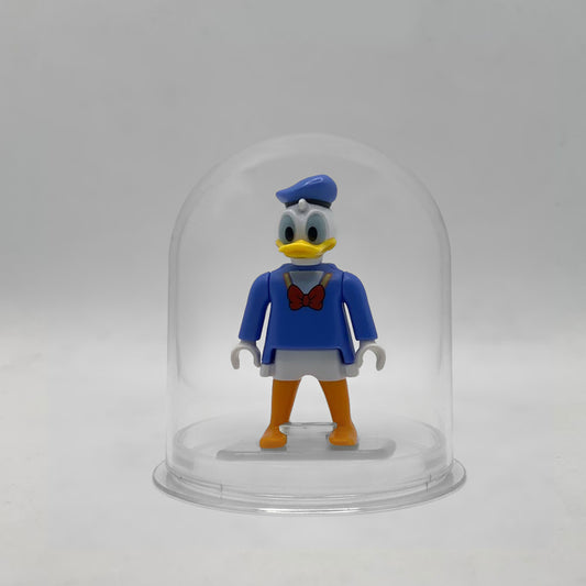 Minifigura Playmobil del Pato Donald con gorra de marinero azul, casaca con pajarita roja y pies naranjas, sobre su peana original Playmobil dentro de la urna expositora transparente, colección Disney Kinder FCO1010