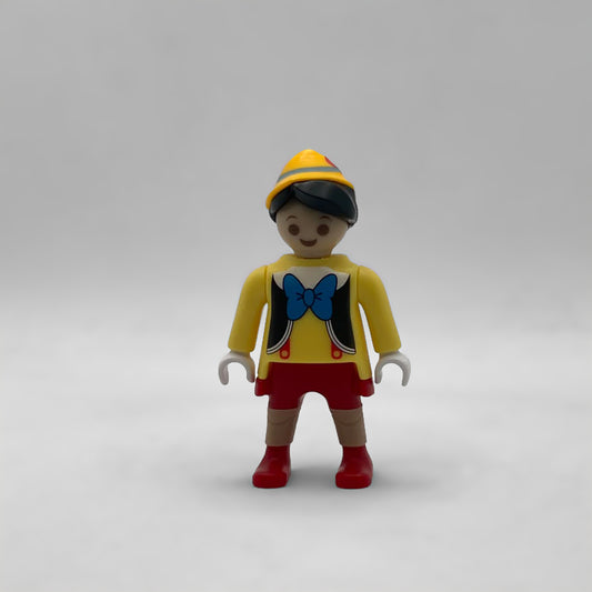 Minifigura Playmobil de Pinocho con gorro amarillo, pajarita azul, tirantes rojos y rodillas de madera moldeadas, colección Disney Kinder FCO1002