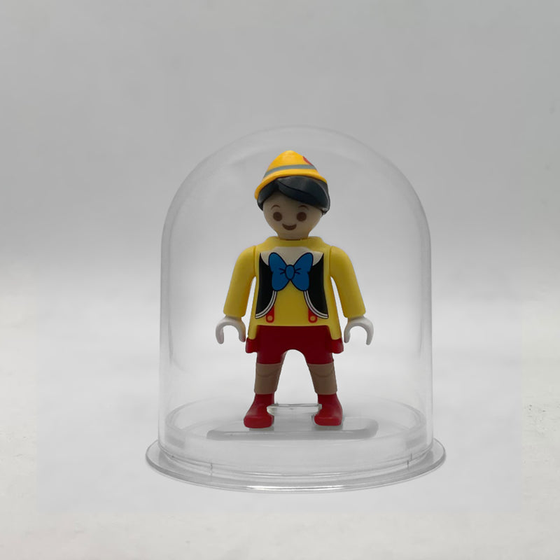 Minifigura Playmobil de Pinocho con gorro amarillo, pajarita azul y rodillas de madera moldeadas, sobre su peana original Playmobil dentro de la urna expositora transparente, colección Disney Kinder FCO1012