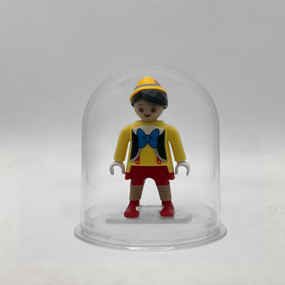 Minifigura Playmobil de Pinocho con gorro amarillo, pajarita azul y rodillas de madera moldeadas, sobre su peana original Playmobil dentro de la urna expositora transparente, colección Disney Kinder FCO1012