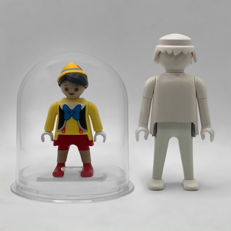 Comparativa de tamaño: Pinocho FCO1012 dentro de su urna expositora junto a una figura Playmobil estándar blanca, que no está incluida
