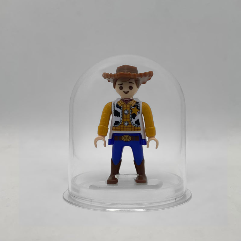 Minifigura Playmobil de Woody con sombrero vaquero, chaleco de vaca y estrella de sheriff, sobre su peana original Playmobil dentro de la urna expositora transparente, colección Disney Kinder FCO1013